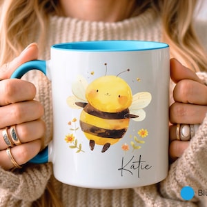 May include: White ceramic mug with a blue interior and handle, featuring a cartoon bee illustration. The bee is yellow and black, with floral accents and the name "Katie" in script. The mug is held by a person.