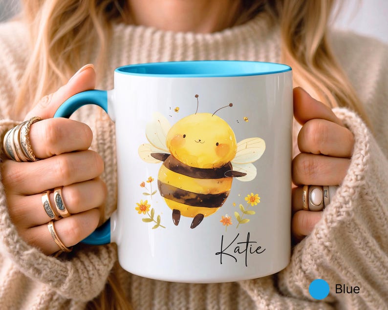 May include: White ceramic mug with a blue interior and handle, featuring a cartoon bee illustration. The bee is yellow and black, with floral accents and the name "Katie" in script. The mug is held by a person.