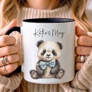 Könnte beinhalten: Weiße Keramiktasse mit schwarzem Rand und Henkel. Die Tasse zeigt eine Illustration eines schwarz-weißen Pandabären mit einer blauen Fliege und dem Text "Katie's Mug" auf der Vorderseite.