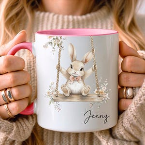 May include: A white ceramic mug with a pink rim features a watercolor illustration of a bunny rabbit on a swing with floral accents. The mug has the name "Jenny" written in cursive at the bottom.