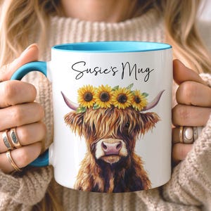 May include: White ceramic mug with a blue interior and handle, featuring a watercolor illustration of a Highland cow wearing a sunflower crown. The mug has the text "Susie's Mug" in black script. A charming and unique gift idea.