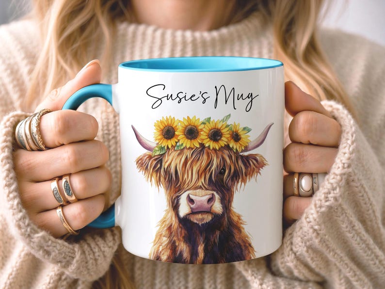 May include: White ceramic mug with a blue interior and handle, featuring a watercolor illustration of a Highland cow wearing a sunflower crown. The mug has the text "Susie's Mug" in black script. A charming and unique gift idea.