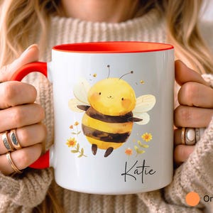 May include: White ceramic mug with a red interior and handle. The mug features a cartoon bee illustration with yellow, black, and white colors, along with the name "Katie" in script. The mug is decorated with small yellow flowers.