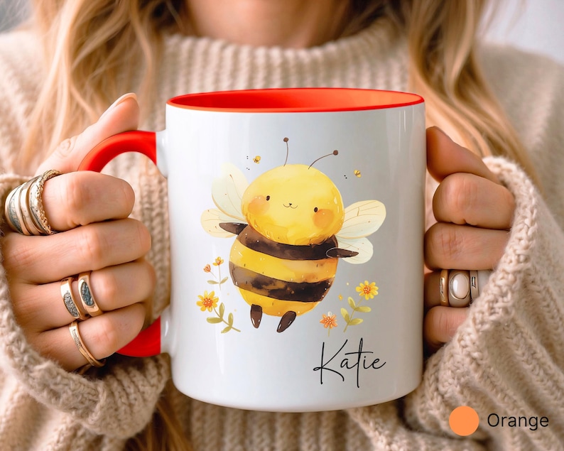 May include: White ceramic mug with a red interior and handle. The mug features a cartoon bee illustration with yellow, black, and white colors, along with the name "Katie" in script. The mug is decorated with small yellow flowers.