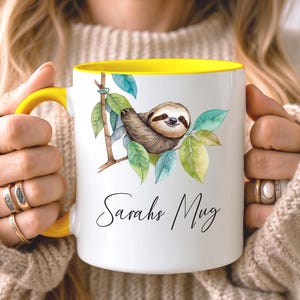 May include: White ceramic mug with a yellow interior and handle. The mug features a watercolor illustration of a sloth on a branch with green leaves. The text "Sarahs Mug" is written in a script font.