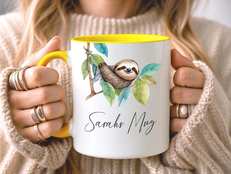May include: White ceramic mug with a yellow interior and handle. The mug features a watercolor illustration of a sloth on a branch with green leaves. The text "Sarahs Mug" is written in a script font.