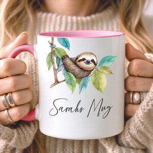 May include: White ceramic mug with a pink interior and handle. The mug features a watercolor illustration of a sloth on a branch with green and blue leaves. The text "Sarahs Mug" is written in a script font.