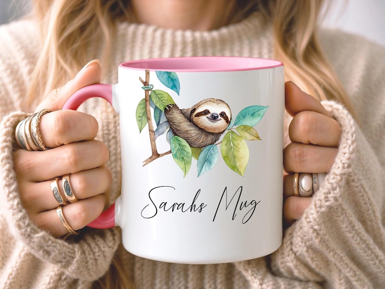May include: White ceramic mug with a pink interior and handle. The mug features a watercolor illustration of a sloth on a branch with green and blue leaves. The text "Sarahs Mug" is written in a script font.