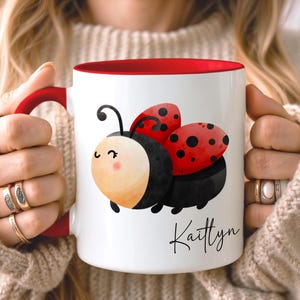 May include: White ceramic mug with a red interior and handle, featuring a cartoon ladybird design. The ladybird has red wings with black spots and a smiling face. The name "Kaitlyn" is written in script.