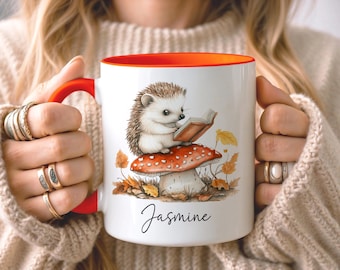 May include: A white ceramic mug with a bright orange handle and interior rim. The mug features a graphic of a small hedgehog sitting on a red and white spotted mushroom while reading a book, surrounded by autumn leaves and the name Jasmine.