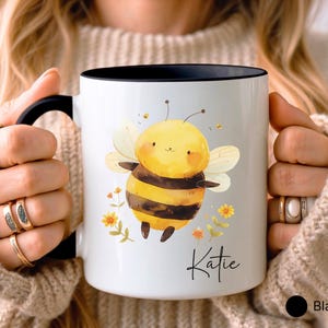 May include: White ceramic mug with a black interior and handle. The mug features a cartoon bee illustration with yellow and black stripes, wings, and floral accents. The name "Katie" is written in script.