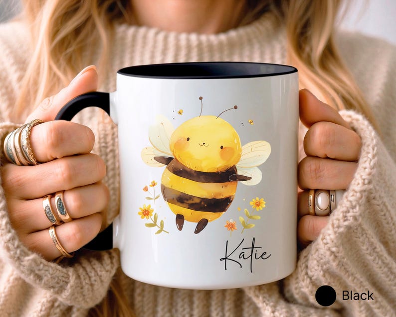 May include: White ceramic mug with a black interior and handle. The mug features a cartoon bee illustration with yellow and black stripes, wings, and floral accents. The name "Katie" is written in script.