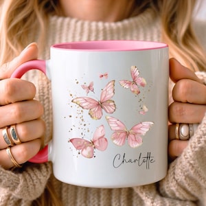Peut inclure: Un mug en céramique blanche avec un intérieur et une anse roses, orné d'un motif aquarelle représentant plusieurs papillons roses et des taches dorées. Le prénom Charlotte est imprimé en police cursive noire sur le côté inférieur droit.