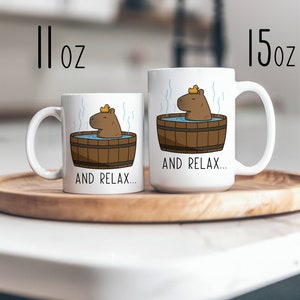 May include: Two white ceramic mugs with a cartoon capybara wearing a yellow rubber ducky hat and relaxing in a wooden hot tub. The text "AND RELAX..." is printed on both mugs. The mug on the left is 11 oz and the mug on the right is 15 oz.