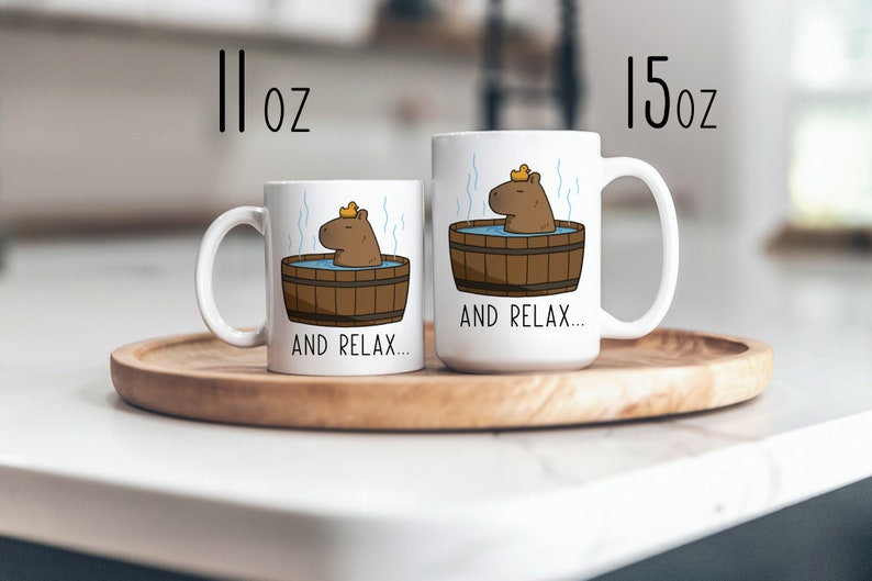 May include: Two white ceramic mugs with a cartoon capybara wearing a yellow rubber ducky hat and relaxing in a wooden hot tub. The text "AND RELAX..." is printed on both mugs. The mug on the left is 11 oz and the mug on the right is 15 oz.