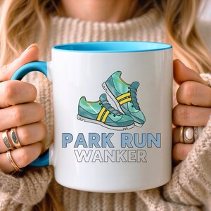 Könnte beinhalten: Weiße Keramiktasse mit blauem Rand und Henkel. Die Tasse zeigt eine Grafik mit zwei grün-blauen Laufschuhen mit gelben Akzenten. Der Text "PARK RUN WANKER" ist in Schwarz unter den Schuhen aufgedruckt.