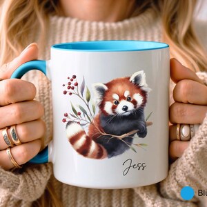 Puede incluir: Taza de cer&aacute;mica blanca con borde y asa azules. La taza presenta una ilustraci&oacute;n de acuarela de un panda rojo sosteniendo una rama con bayas. El nombre "Jess" est&aacute; escrito en cursiva en la parte inferior de la taza.