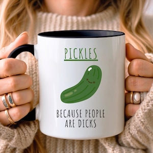 Op de afbeelding: Een witte keramische mok met een zwarte handgreep en binnenrand. De voorzijde toont een groene cartoon augurk met de tekst PICKLES erboven en de tekst BECAUSE PEOPLE ARE DICKS in zwarte letters onder de afbeelding van de augurk.