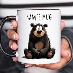 May include: A white ceramic mug with a black handle and rim, featuring a cartoon illustration of a sitting black bear with a light brown belly and the text "SAM'S MUG" printed in black lettering above the bear on the front of the mug.