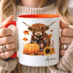 May include: A white ceramic mug with a bright orange handle and interior rim. The mug features a graphic of a brown Highland cow wearing a plaid scarf, sitting next to a large orange pumpkin and a sunflower, surrounded by autumn leaves and the name Anna.