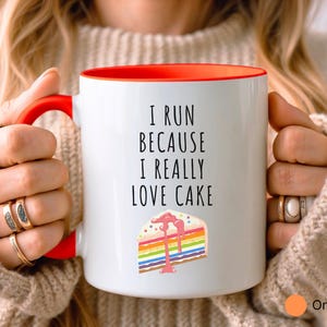 May include: White coffee mug with an orange rim. The mug has a black inscription that reads "I run because I really love cake". A colorful cake slice with rainbow frosting is pictured below the text.