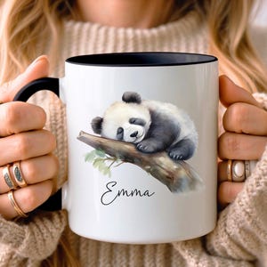 Könnte beinhalten: Weiße Keramiktasse mit schwarzem Rand und Henkel. Die Tasse zeigt eine Aquarell-Illustration eines schlafenden Pandabären auf einem Ast mit grünen Blättern. Der Name "Emma" ist unter dem Panda in Kursivschrift geschrieben.