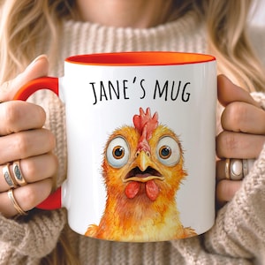 Op de afbeelding: Een witte keramische mok met een feloranje handvat en binnenrand. De voorzijde toont een kleurrijke aquarelillustratie van een kip met grote ogen en oranje en gele veren. De tekst JANE'S MUG is in het zwart boven de kip gedrukt.