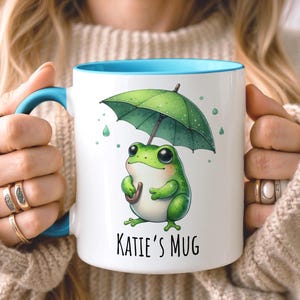 Könnte beinhalten: Weiße Keramik-Tasse mit blauem Innenraum und Griff. Die Tasse zeigt einen grünen Frosch, der einen grünen Regenschirm hält, mit dem Text "Katie's Mug" darunter. Die Tasse wird von einer Person gehalten.