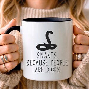 Könnte beinhalten: Ein weißer Keramikbecher mit schwarzem Innenraum und Henkel. Auf der Vorderseite befindet sich eine schwarze Grafik einer zusammengerollten Schlange über dem schwarzen Text: SNAKES. BECAUSE PEOPLE ARE DICKS. Der Becher wird von zwei Händen mit goldenen Ringen gehalten.