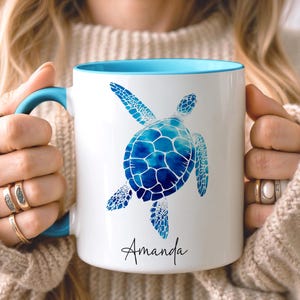May include: White ceramic mug with a blue interior and handle. The mug features a blue watercolor sea turtle design. The name "Amanda" is written in a script font below the turtle.