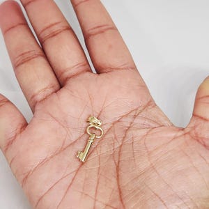 May include: A small, gold-colored key pendant. The key has a classic design with a bow, stem, and bit. The pendant is likely made of gold and is designed to be worn as jewelry. The key is shown against a neutral background.