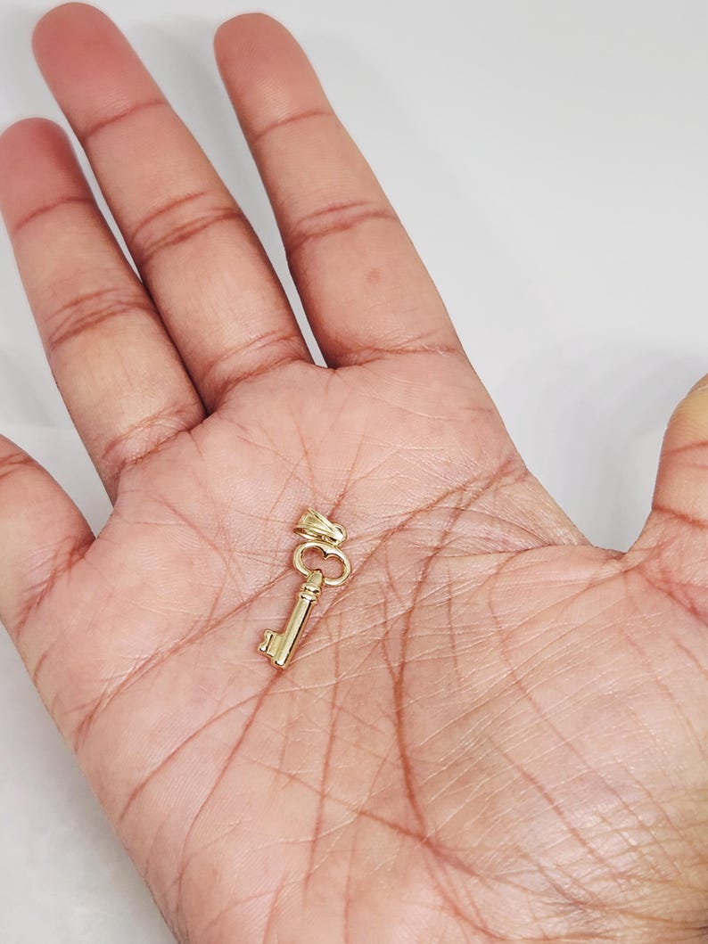 May include: A small, gold-colored key pendant. The key has a classic design with a bow, stem, and bit. The pendant is likely made of gold and is designed to be worn as jewelry. The key is shown against a neutral background.