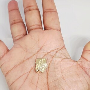 May include: A gold pendant shaped like a small fan of four playing cards. The cards are engraved with suit symbols including a spade, heart, diamond, and club. A small circular bail is attached to the bottom corner for hanging on a chain.