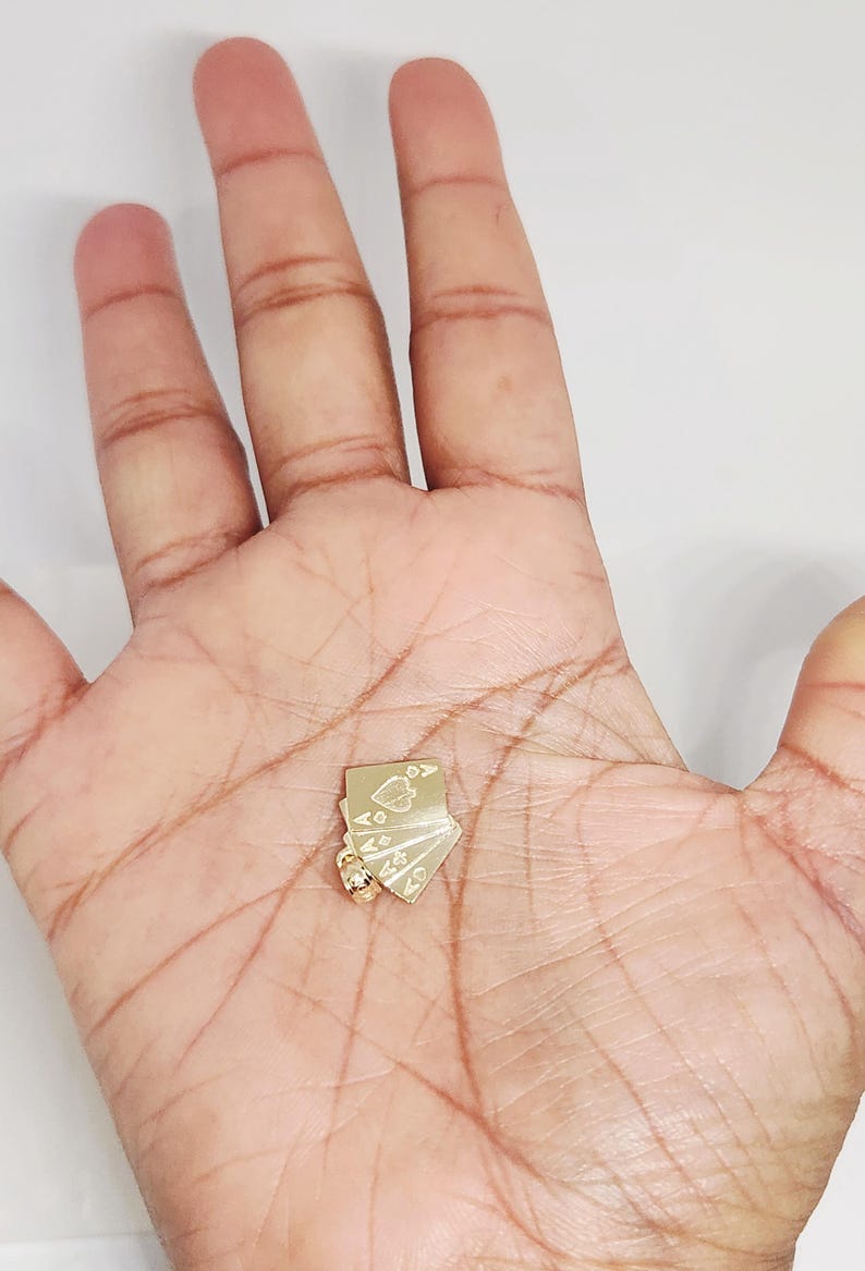 May include: A gold pendant shaped like a small fan of four playing cards. The cards are engraved with suit symbols including a spade, heart, diamond, and club. A small circular bail is attached to the bottom corner for hanging on a chain.