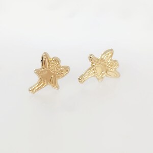 Puede incluir: Un par de pendientes de bot&oacute;n dorados con forma de hada. Cada pendiente presenta un dise&ntilde;o detallado de hada con alas extendidas y un vestido fluido. Los pendientes est&aacute;n sobre un fondo blanco.