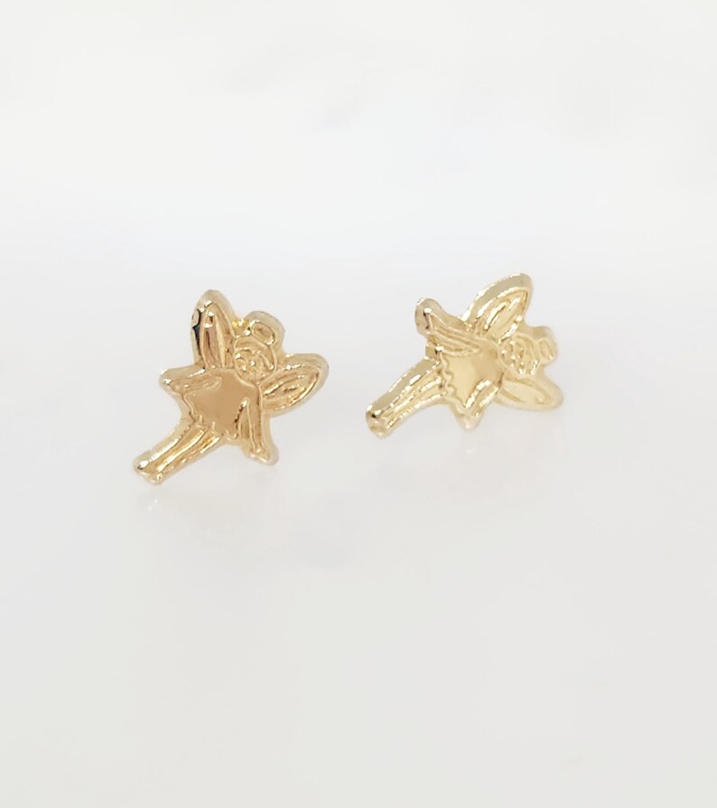 Puede incluir: Un par de pendientes de bot&oacute;n dorados con forma de hada. Cada pendiente presenta un dise&ntilde;o detallado de hada con alas extendidas y un vestido fluido. Los pendientes est&aacute;n sobre un fondo blanco.