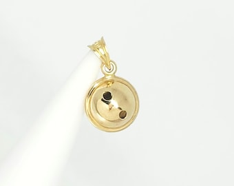 May include: A polished gold pendant shaped like a small bowling ball. The spherical charm features two dark circular indentations on its front surface and includes a small triangular bail at the top for attachment to a necklace chain.