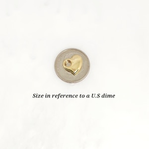 May include: A small, polished gold-tone heart charm featuring a circular loop at the top for attachment. The charm is positioned on top of a U.S. dime to provide a clear visual reference for its size, which is approximately 1.79 cm in diameter.
