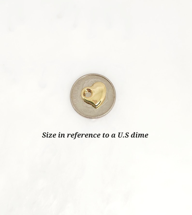 May include: A small, polished gold-tone heart charm featuring a circular loop at the top for attachment. The charm is positioned on top of a U.S. dime to provide a clear visual reference for its size, which is approximately 1.79 cm in diameter.