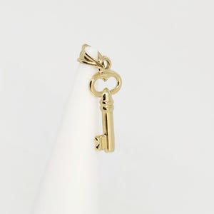 May include: A gold-colored key pendant with a heart-shaped design on the top and a detailed key shaft. The pendant is suspended from a small loop, and the key is displayed against a white background.