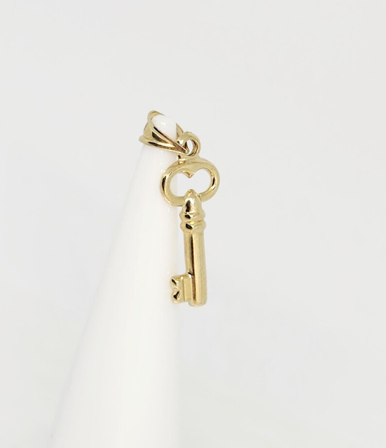 May include: A gold-colored key pendant with a heart-shaped design on the top and a detailed key shaft. The pendant is suspended from a small loop, and the key is displayed against a white background.