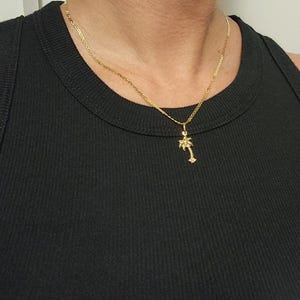 Op de afbeelding: Een goudkleurige ketting met een fijne schakel en een kleine palmboomhanger. De hanger hangt aan een eenvoudig ringetje en bevindt zich centraal tegen een zwarte, geribbelde stof, wat zorgt voor een duidelijk en helder contrast.