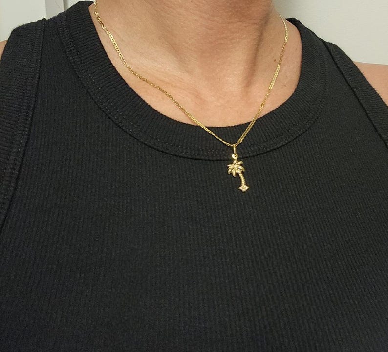 Op de afbeelding: Een goudkleurige ketting met een fijne schakel en een kleine palmboomhanger. De hanger hangt aan een eenvoudig ringetje en bevindt zich centraal tegen een zwarte, geribbelde stof, wat zorgt voor een duidelijk en helder contrast.