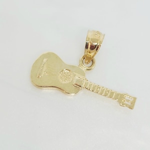 Può includere: Un ciondolo a forma di chitarra color oro. Il ciondolo presenta un design dettagliato di chitarra acustica in miniatura, tra cui corde, buca e tastiera. L'anello è attaccato alla parte superiore della chitarra. Il ciondolo è probabilmente in oro.