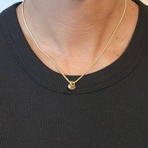 May include: A gold-toned chain necklace featuring a small, round, textured pendant. The necklace rests against a black ribbed vest, showcasing a simple link design and a polished metallic finish that catches the light around the neck area.