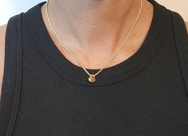 May include: A gold-toned chain necklace featuring a small, round, textured pendant. The necklace rests against a black ribbed vest, showcasing a simple link design and a polished metallic finish that catches the light around the neck area.