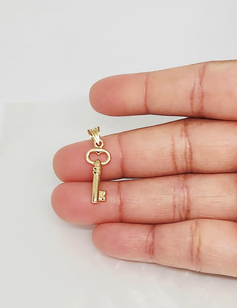 May include: A small, gold-colored key pendant. The key has a detailed design with a loop at the top and a rectangular key bit. The pendant is held against a neutral background.