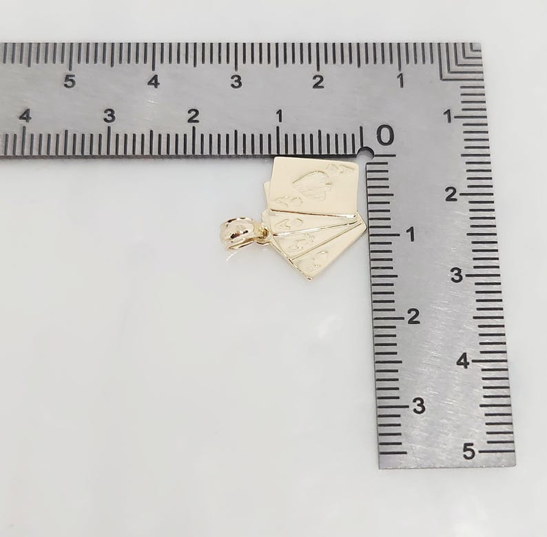 May include: A gold-colored metal pendant shaped like a fan of playing cards, featuring an ace of spades design. The pendant is positioned next to a metal ruler, showing a width of approximately 0.75 inches and a height of 0.75 inches.