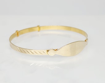 Puede incluir: Una pulsera ajustable de color dorado con una placa ovalada. La pulsera tiene un diseño sencillo con una banda texturizada en un lado y un acabado liso y pulido en la placa de identificación. La pulsera es probablemente de oro o metal chapado en oro.
