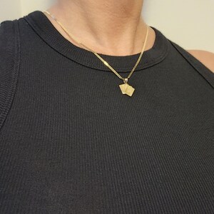 May include: A gold-toned necklace featuring a delicate chain and a small pendant shaped like two playing cards. The pendant hangs from a simple bail, and the entire piece is worn against a black ribbed sleeveless top, highlighting the metallic finish.